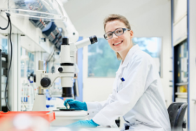 Researcher in white lab coat smiles while sitting in lab
