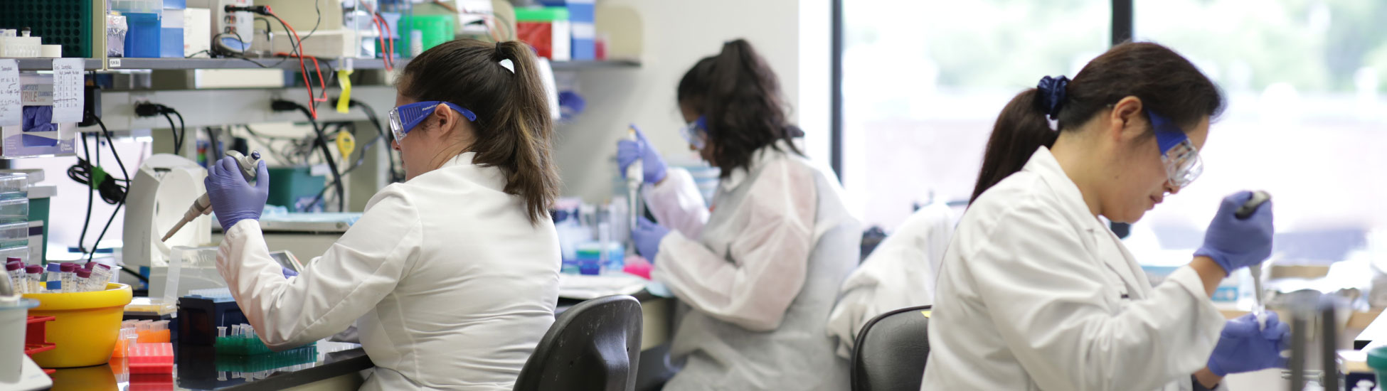 Three UCB researchers in white lab coats sitting in a science lab holding biomedical research tools